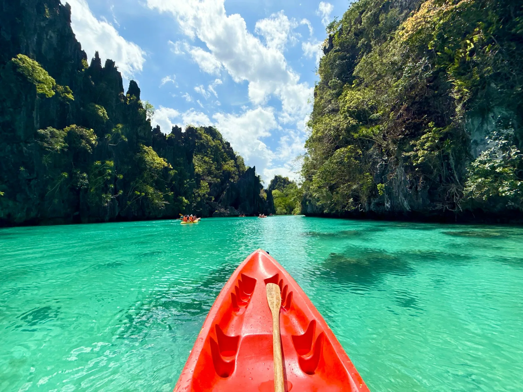 An orange kayak pointing across clear turquoise water toward forested cliffs.